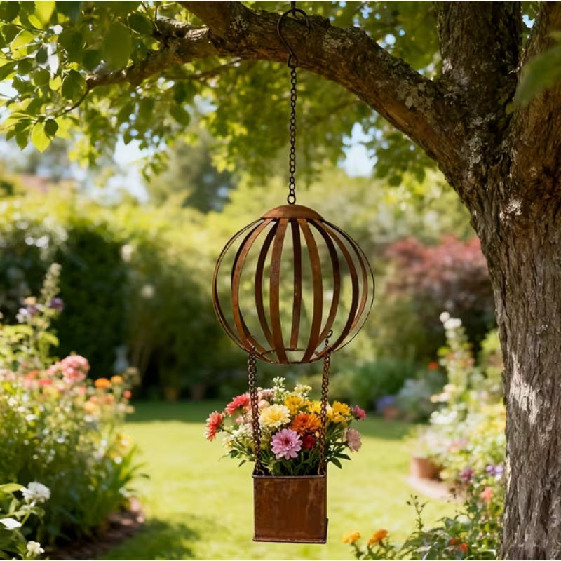 A hanging metal garden sculpture shaped like a hot air balloon, with a rusted copper-toned spherical cage and a matching rectangular pot holding a colorful bouquet of flowers, suspended under a tree in a sunny garden.