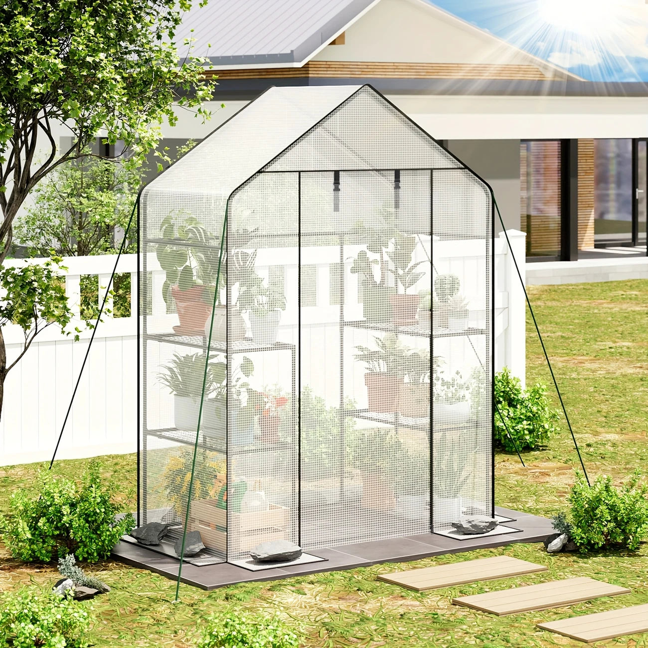 A portable greenhouse with transparent panels set on a wooden base, featuring a white frame and shelves with plants inside. The greenhouse is outdoors, with trees and a wooden fence in the background.