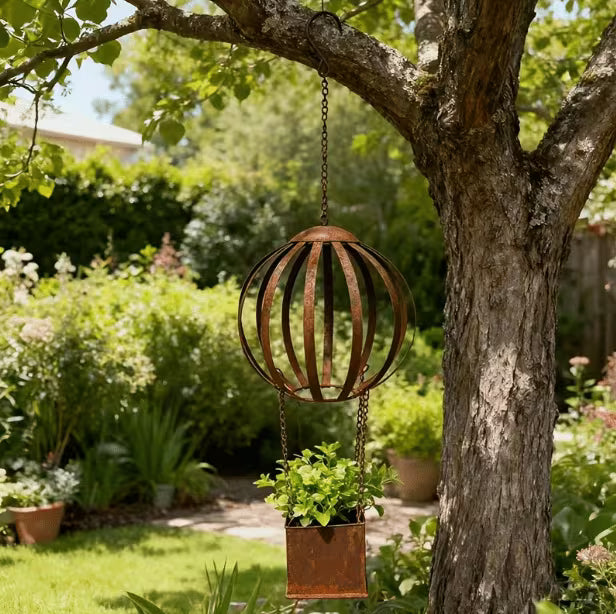 A rusted metal hanging garden sphere with a rectangular planter box suspended from chains, displayed outdoors on a tree in a lush garden.