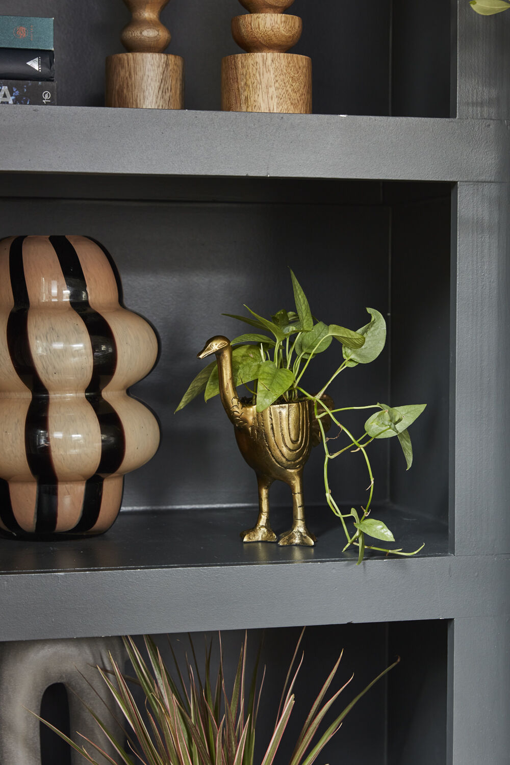 Decorative plant in a gold ostrich planter on a dark shelf with wooden items above.